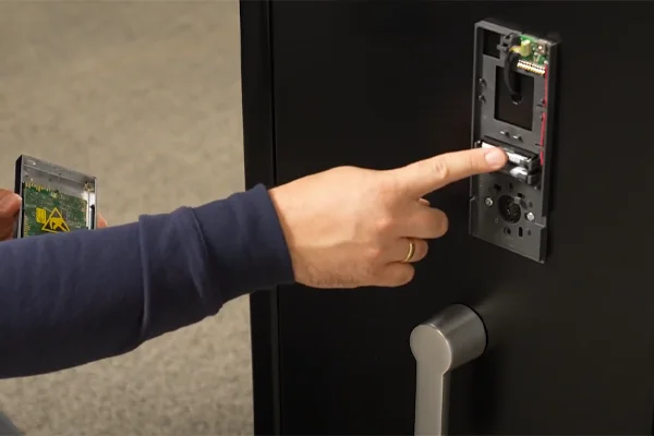 Hand zeigt auf das geöffnete Gehäuse eines elektronischen Tresorschlosses an einer schwarzen Tür, in der anderen Hand die Platine