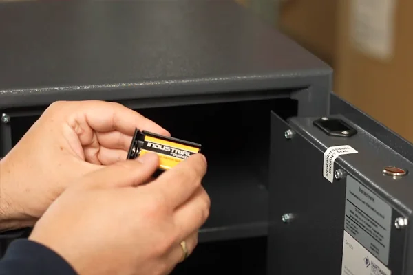 Hände setzen eine 9-Volt-Batterie in das Batteriefach an der Innenseite einer geöffneten Tresortür ein