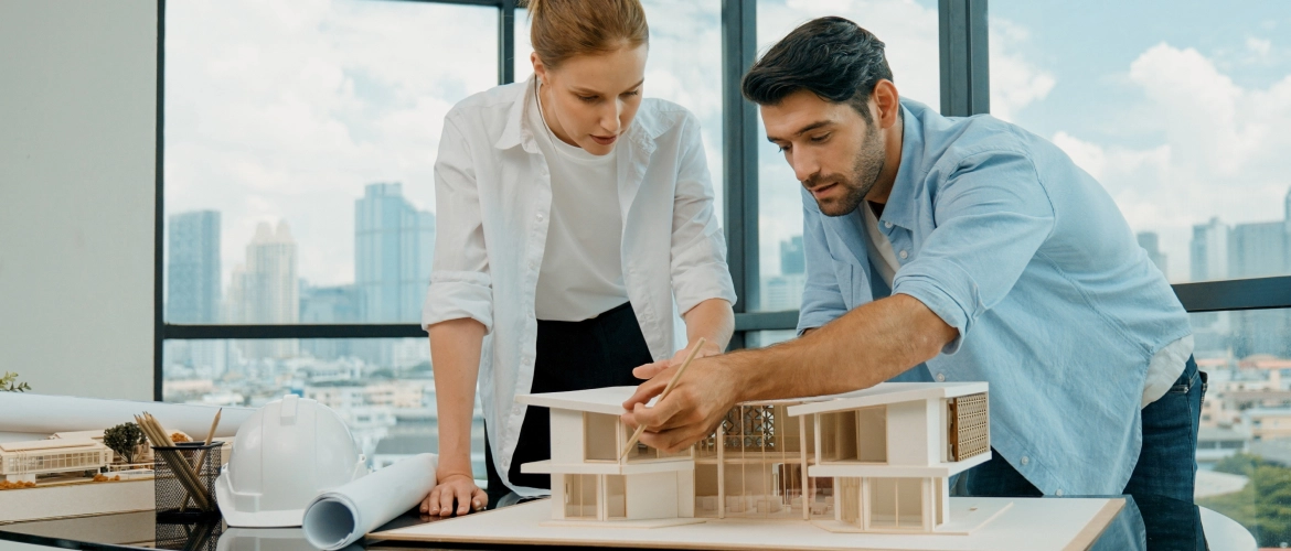 Eine Architektin und ein Architekt besprechen ein Architekturmodell an einem Tisch vor einer Fensterfront mit Stadtblick