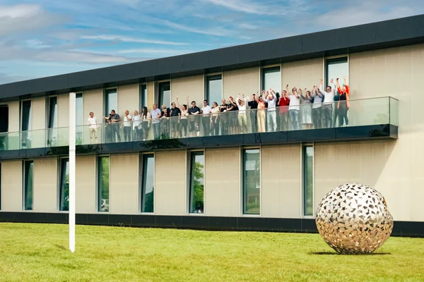Mitarbeitende von HARTMANN TRESORE winken vom Balkon der Firmenzentrale in Paderborn, davor eine Kugelskulptur auf der Wiese