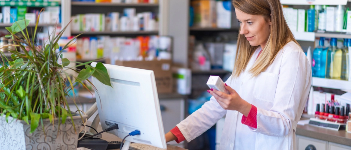 Apothekerin im weißen Kittel mit Medikamentenpackung am Computer, im Hintergrund Regale einer Apotheke