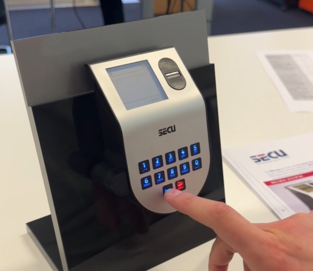 Finger bedient die beleuchtete Tastatur eines SECU-Elektronikschlosses mit Display und Fingerabdruckscanner am Schulungsaufsteller