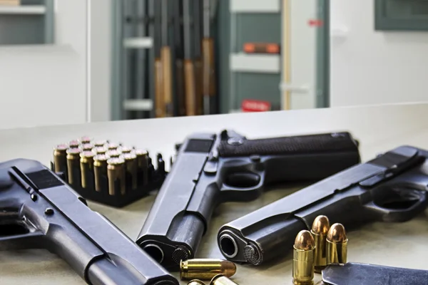 Pistolen und Patronen auf einem Tisch, im Hintergrund ein Waffenschrank mit Langwaffen