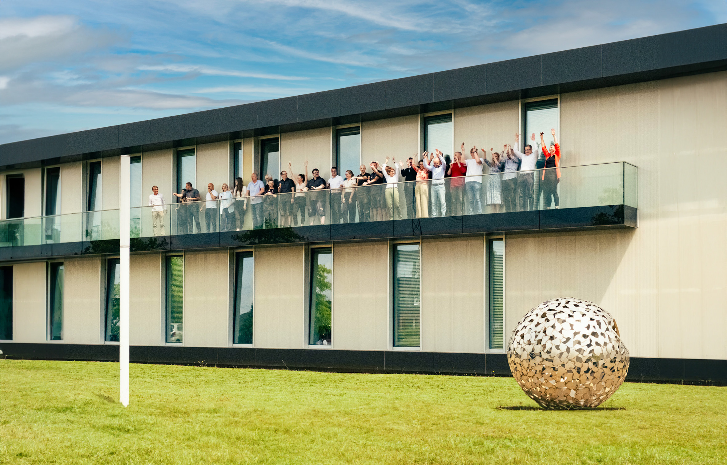 Außenaufnahme des modernen Bürogebäudes der Hartmann Tresore AG mit Mitarbeitenden auf dem Balkon; im Vordergrund eine große, kugelförmige Edelstahlinstallation auf der Rasenfläche – Symbol für Innovation und Sicherheit bei Tresorlösungen.