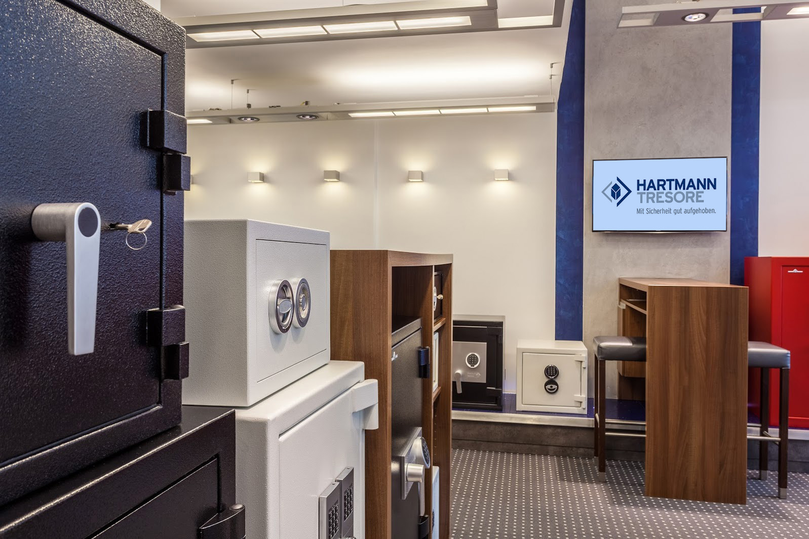 „Moderne Tresorausstellung im Showroom von HARTMANN TRESORE mit verschiedenen Sicherheitstresoren, darunter ein schwarzer Wertschutzschrank mit Schlüsselverriegelung, ein weißer Tresor mit Doppelbartschlössern und mehrere Möbeltresore mit elektronischem Zahlenschloss – perfekt für private und gewerbliche Sicherheitslösungen.“